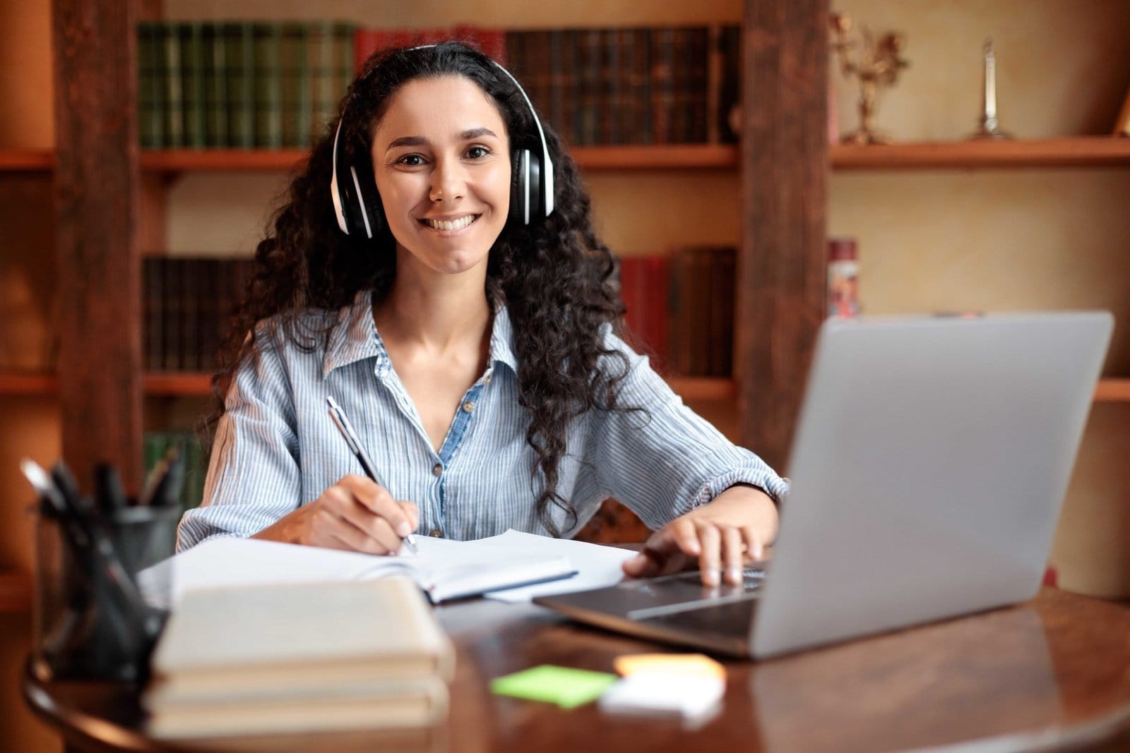 A smiling woman with headphones taking notes at a laptop