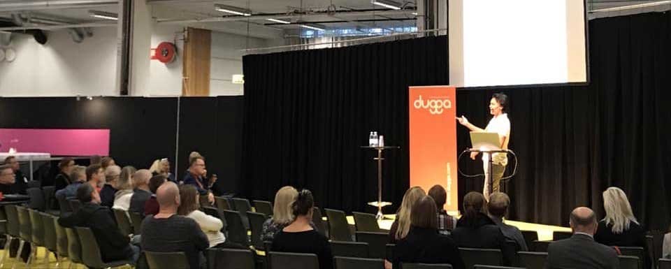 A speaker on stage in front of a Dugga banner, presenting to an audience