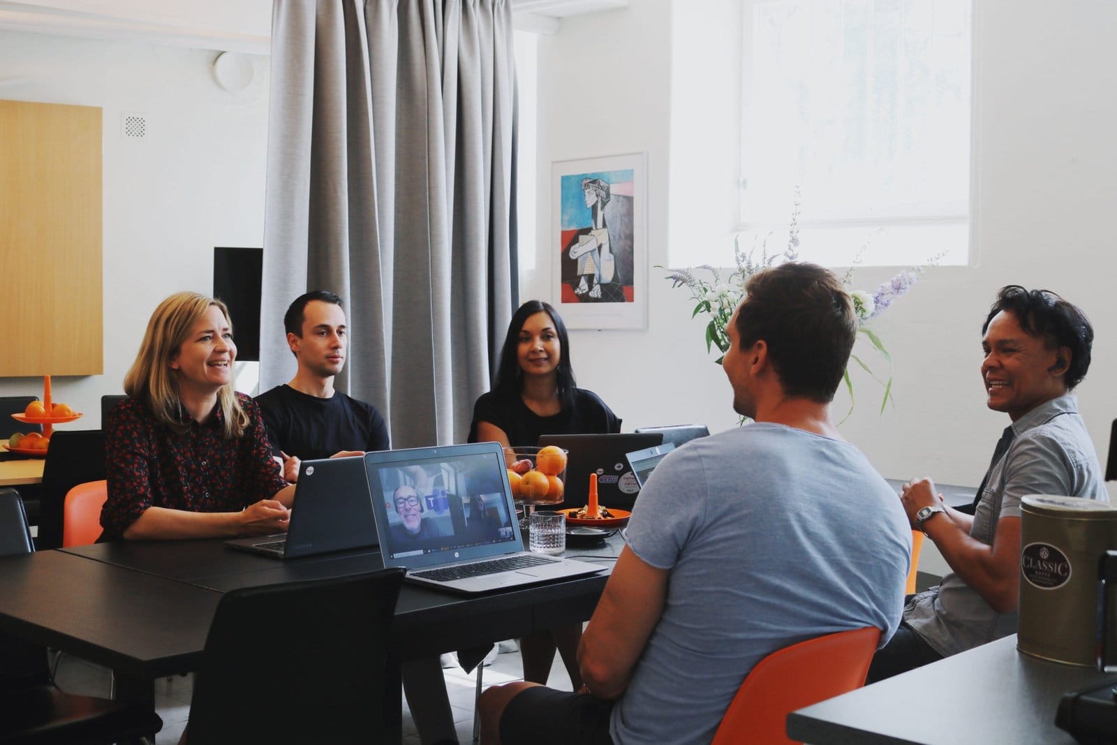 A team meeting around a table, with one colleague joining on a video call